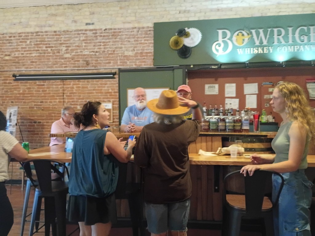 Interior of Bowright Whiskey Company with patrons socializing at the bar, brick walls in background, and various whiskey bottles displayed behind the counter.