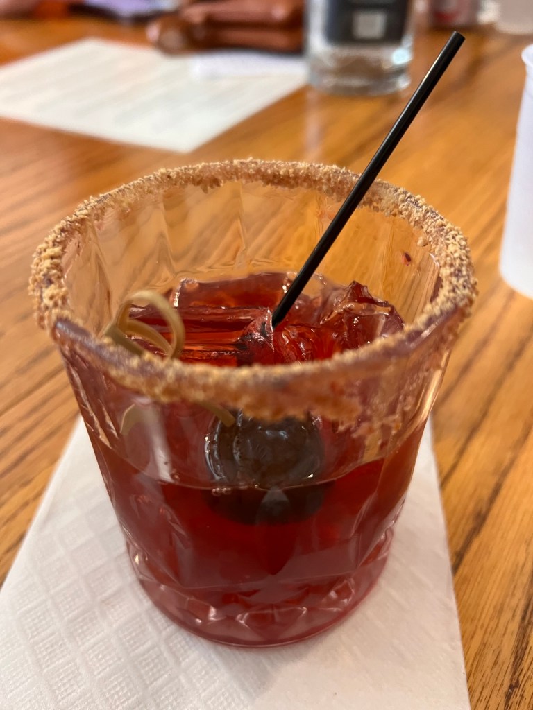 A close-up of a cocktail in a glass with a sugared rim, featuring ice cubes and a cherry, topped with a black straw, placed on a napkin on a wooden table.