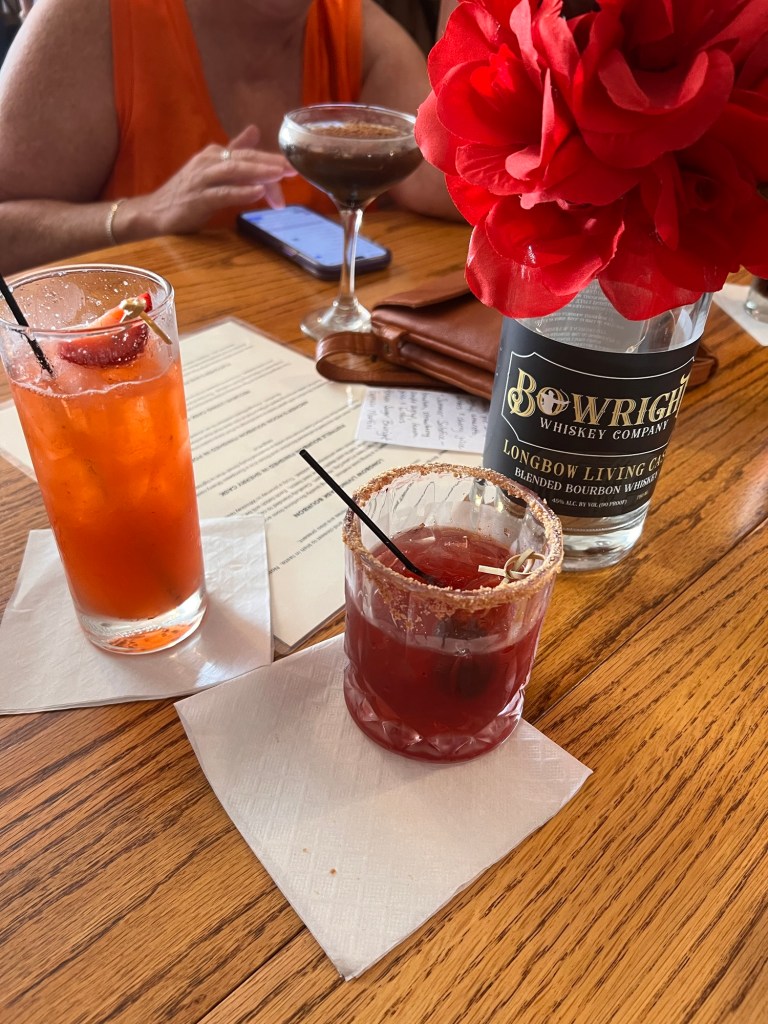 A table setting with two cocktails: one garnished with a strawberry and the other a cherry, with a decorative red flower in the background and menus on the table.