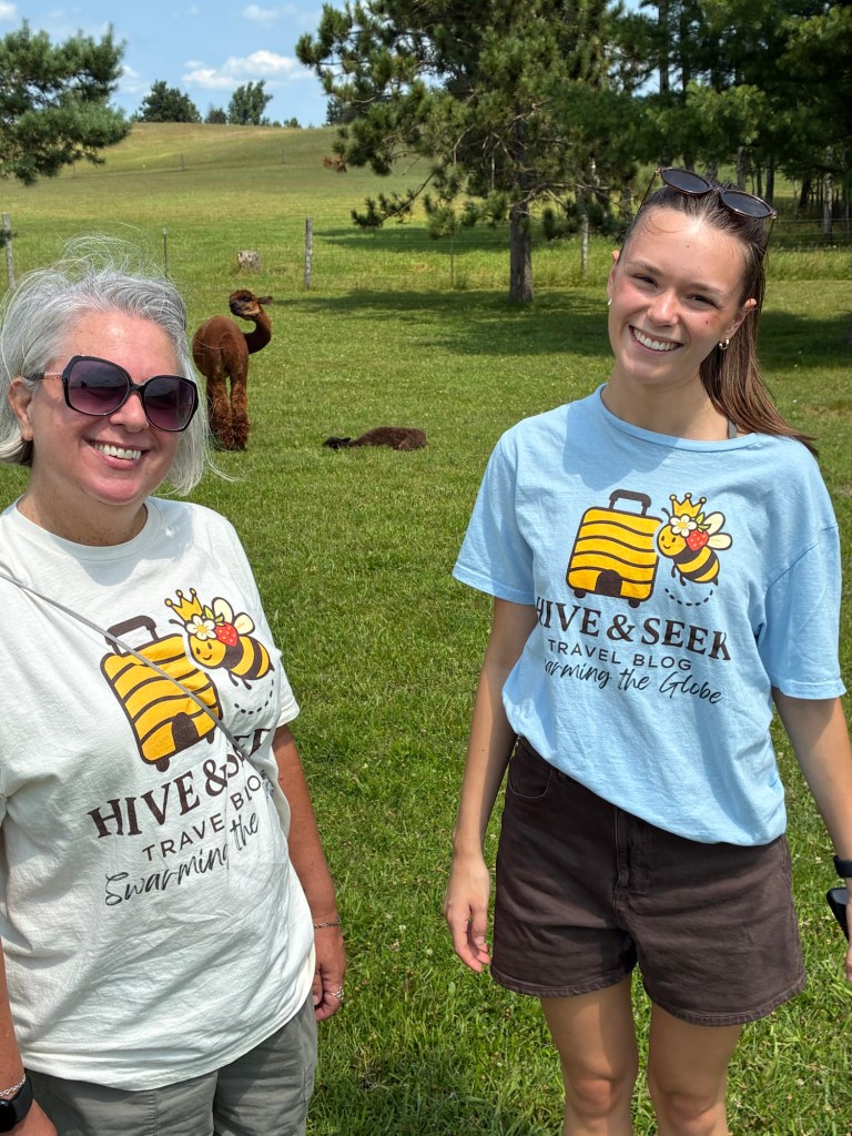 Two women smiling for the camera, wearing matching travel blog T-shirts, standing in a grassy field with alpacas in the background.