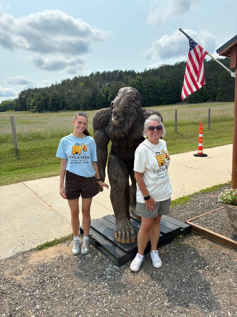 Two women pose next to a large Bigfoot statue outdoors, with an American flag in the background and a field of trees.